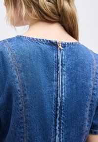 Πίσω όψη ενός ατόμου που φοράει μπλε τοπ από τζιν με κοντό φερμουάρ στο πάνω μέρος της πλάτης και ορατές ραφές.