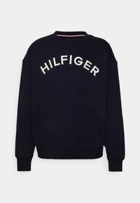 Sudadera azul marino con cuello redondo, que presenta el texto "HILFIGER" bordado en blanco en un diseño arqueado. Tela suave, con puños y el dobladillo acanalados.