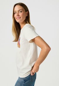 Crèmekleurig katoenen T-shirt met een kleurrijk grafisch ontwerp op de voorkant. Korte mouwen en een relaxte pasvorm, gecombineerd met blauwe jeans.