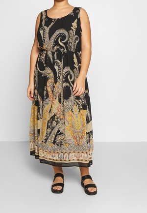 Frau trägt ein schwarzes ärmelloses Maxikleid mit filigranen goldenen und beigen Paisley-Mustern und schwarzen Sandalen mit Knöchelriemchen.