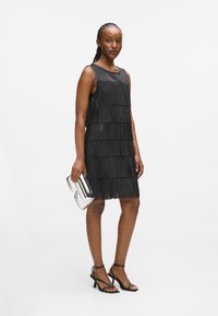 Μαύρο φόρεμα με φούντες και στρωτή υφή, χωρίς μανίκια και στρογγυλή λαιμόκοψη. Συνδυασμένο με ασημί γεωμετρική τσάντα και μαύρα πέδιλα με λουράκια.