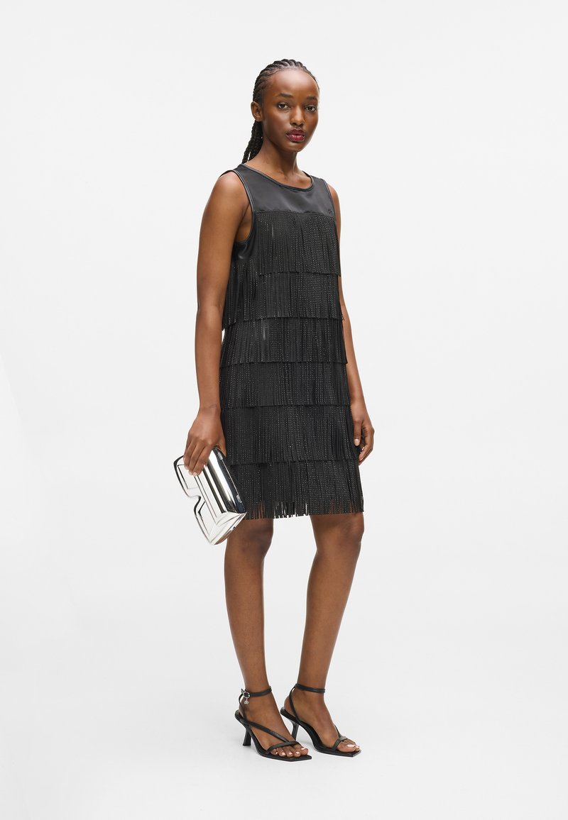 Μαύρο φόρεμα με φούντες και στ�ρωτή υφή, χωρίς μανίκια και στρογγυλή λαιμόκοψη. Συνδυασμένο με ασημί γεωμετρική τσάντα και μαύρα πέδιλα με λουράκια.