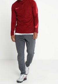 Chemise de sport rouge à manches longues avec demi-fermeture éclair et accents texturés sur les côtés, associée à un pantalon de survêtement gris et des chaussures de sport grises.