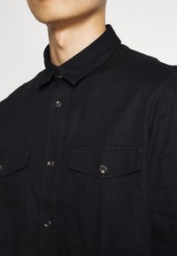 Camisa negra de botones con cuello clásico, tejido texturizado, dos bolsillos en el pecho y botones redondos. Vista en primer plano de la sección superior.