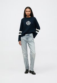 Pulover tricotat navy cu model texturat și logo alb, asortat cu blugi bleu deschis, drepți, și pantofi negri cu vârful rotunjit.