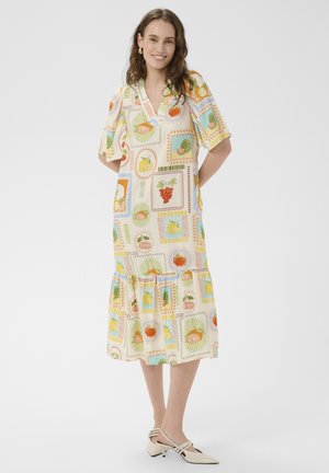 Žena sa vlnitými vlasmi, ktorá sa usmieva, nosi midi šaty s farebnými ilustráciami ovocia a béžovými nízkymi opätkami na bielom pozadí.