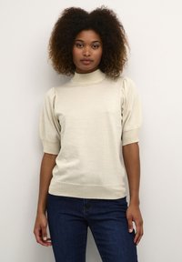 Haut beige clair à manches courtes avec un col haut, en tissu tricoté texturé et subtilement scintillant, associé à un jean bleu foncé.
