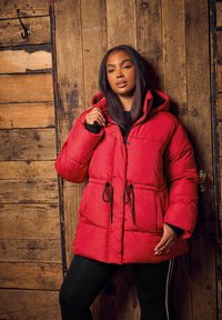 Doudoune rouge avec une taille élastique, poches avant et capuche. Présente une texture matelassée et des accents noirs aux poignets.