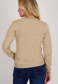 Beige langemouwtop in ribbeltextuur met ronde hals en aansluitende manchetten, gecombineerd met blauwe denim jeans met achterzakken.
