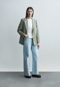 Žena stojí oblečená v zelenom pruhovanom blejzri, bielej nariasenej blúzke, svetlomodrých širokých džínsoch a hnedých platformových topánkach na sivom podlahe.