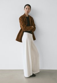 Femme aux cheveux noirs attachés, portant une veste en daim marron, un haut noir, un pantalon large blanc et des chaussures noires, debout les bras croisés.