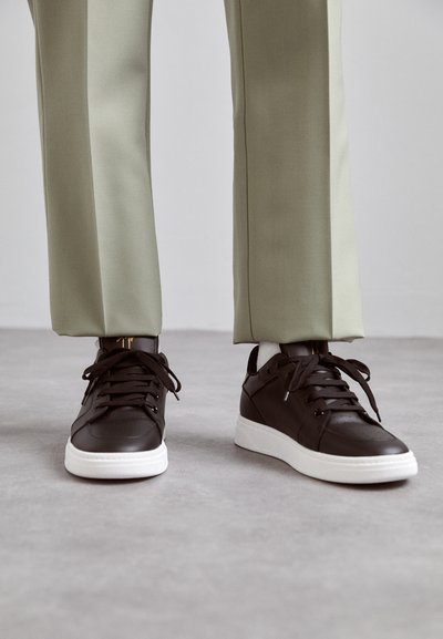 Brune lædersneakers med perforerede detaljer, flad hvid gummisål og sorte snørebånd, sat sammen med lysegrønne tapered bukser.