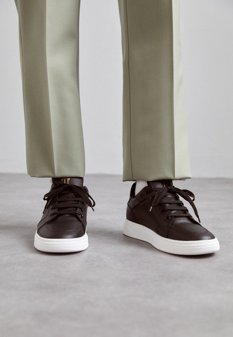 Adidași din piele maro cu detalii perforate, talpă plată din cauciuc alb și șireturi negre, asortați cu pantaloni ușor verzi, tapered.