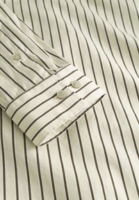 Cămașă în dungi, dintr-un material crem deschis, cu dungi subțiri maro închis. Are manșetă cu nasturi, cu două nasturi și detalii vizibile de cusătură.