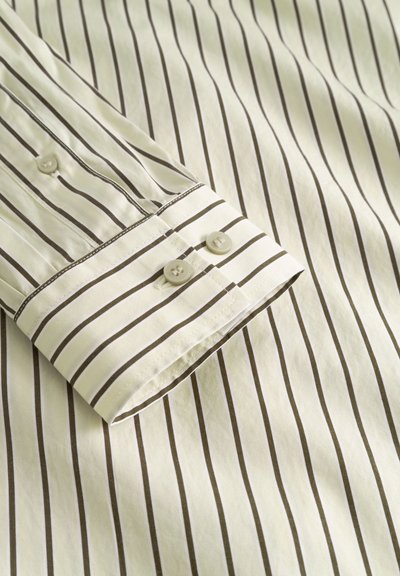 Chemise rayée en tissu crème clair avec de fines rayures brun foncé. Elle possède un poignet boutonné avec deux boutons et des détails de surpiqûres visibles.