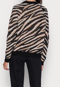 Person som bär en stickad tröja med svarta, beige och tan zebraränder, ihop med svarta byxor, sedd bakifrån mot en vit bakgrund.