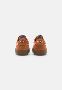 Nike sportcipő narancssárga velúr felsőrésszel, fehér részletekkel és barna gumitalppal. A márkaneve a saroknál látható.