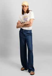 Căciulă bej, tricou alb cu design floral și blugi largi de culoare albastru închis. Sandale marron completează ținuta. Look simplu, casual.