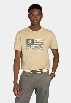 Joven con camiseta gráfica beige con bandera camuflada y herramientas cruzadas, pantalones cargo grises, de pie con una mano en el bolsillo.