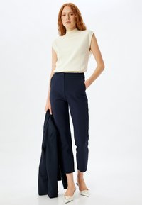 Hvid ærmeløs top, marineblå skræddersyede bukser og hvide spidse pumps. En mørk blazer er draperet over den ene arm. Enkel, ren design.
