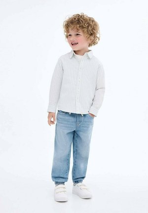 Une chemise à rayures grises claires à manches longues, un pantalon en denim bleu avec des poignets élastiques et des baskets blanches. Un enfant se tient en souriant devant un fond blanc.