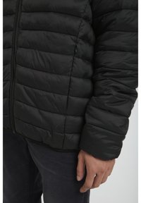 Black padded jacket featuring a quilted texture, zip fastening, and wide cuffs, with a smooth finish and minimalistic design.