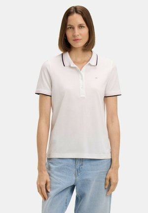 Femme aux cheveux bruns mi-longs portant un polo blanc avec des bordures noires et roses et un jean bleu clair, debout devant un fond uni.