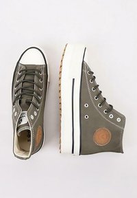 Un par de zapatillas altas verde oliva con suelas de goma blanca, cordones negros y un parche circular marrón en el lado.