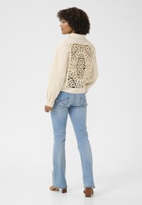 Beige, lätt jacka med en spetscrochet-detalj på ryggen, i kombination med ljusblå utsvängda jeans och bruna klacksandaler.