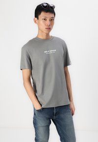 Camiseta de algodón gris con mangas cortas, que presenta "REPLAY GARAGE ESTABLISHED 1981" impreso en blanco, combinada con jeans de denim azul.