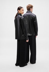 Divas personas valkā melnas ādas oversized kreklus ar gludu tekstūru un pogām aizpogātas aproces, kombinēti ar plašām melnām biksēm.