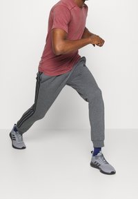 Šedé sportovní tepláky s černými bočními pruhy, v kombinaci s červeným tričkem s krátkým rukávem a šedými teniskami, v dynamické běžecké pozici.