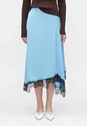 Jupe asymétrique bleue en tissu léger avec un ourlet en dentelle noire. Présente des bords effilochés et une texture lisse. Portée avec des chaussures blanches.