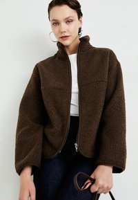 Young woman with short hair wears oversized brown fleece jacket over white top and dark jeans, holding a brown strap in hand.