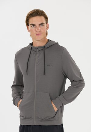 Young man wearing a gray zip-up hoodie with front pockets and black drawstrings, standing with hands in pockets against a white background.