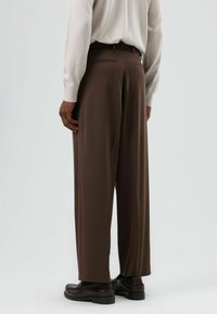 Bruine broek met wijde pijpen, met een gladde textuur en twee achterzakken. Gecombineerd met zwarte schoenen en een lichtgekleurde blouse.