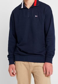 Felpa blu navy con colletto alto, chiusura a zip e dettagli colorati al collo. Realizzata in tessuto morbido, presenta polsini ed orlo elasticizzati.
