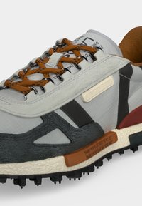 Gros plan d'une basket blanche et grise avec des lacets marron tachetés, une semelle texturée et un logo Lacoste sur le panneau latéral.