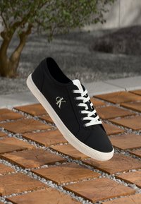 Czarne sneakersy z białymi sznurowadłami i logo "cK", zawieszone nad drewnianymi kwadratowymi płytkami otoczonymi małymi szarymi kamieniami na zewnątrz.