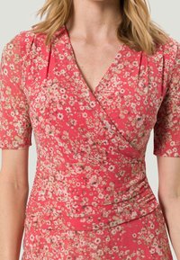 Kukkakuosi mekko, jossa on vaaleanpunainen pohja ja pieniä valkoisia sekä keltaisia kukkia. Lyhyet hihat ja kerätty yksityiskohta vyötäröllä. Pehmeä kankaan rakenne.