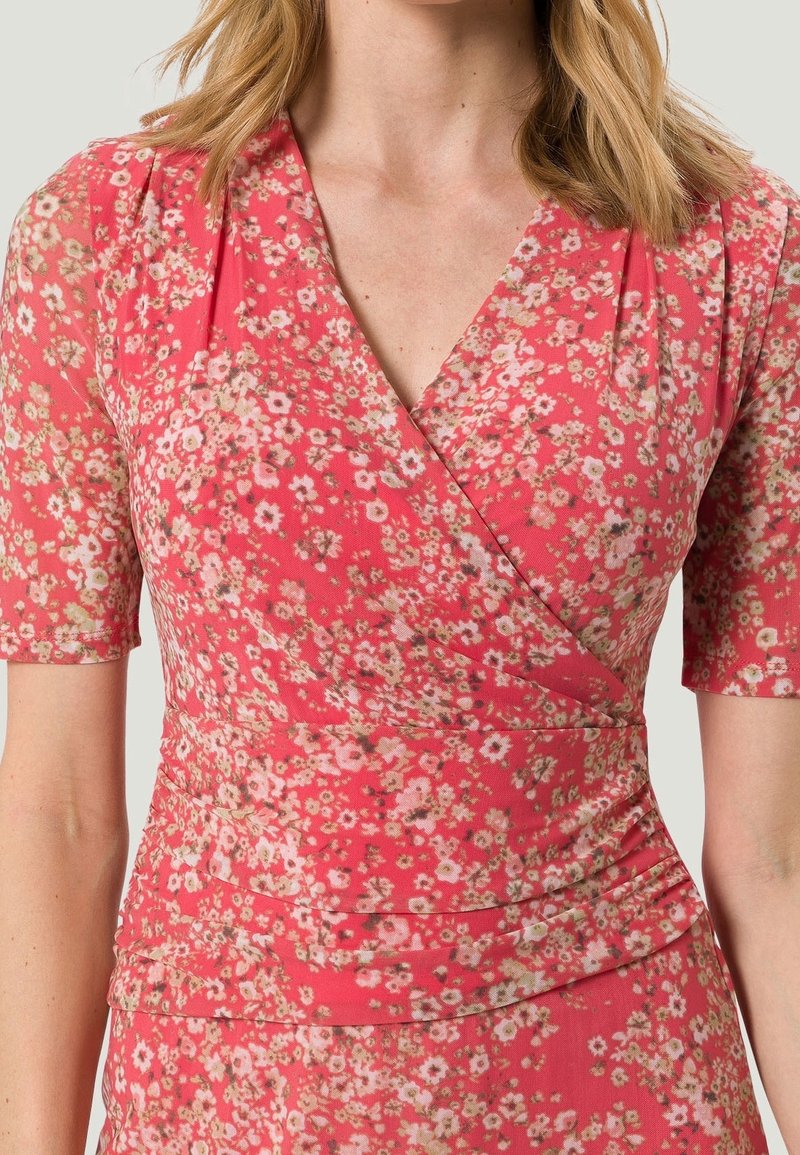 Kukkakuosi mekko, jossa on vaaleanpunainen pohja ja pieniä valkoisia sekä keltaisia kukkia. Lyhyet hihat ja kerätty yksityiskohta vyötäröllä. Pehmeä kankaan rakenne.