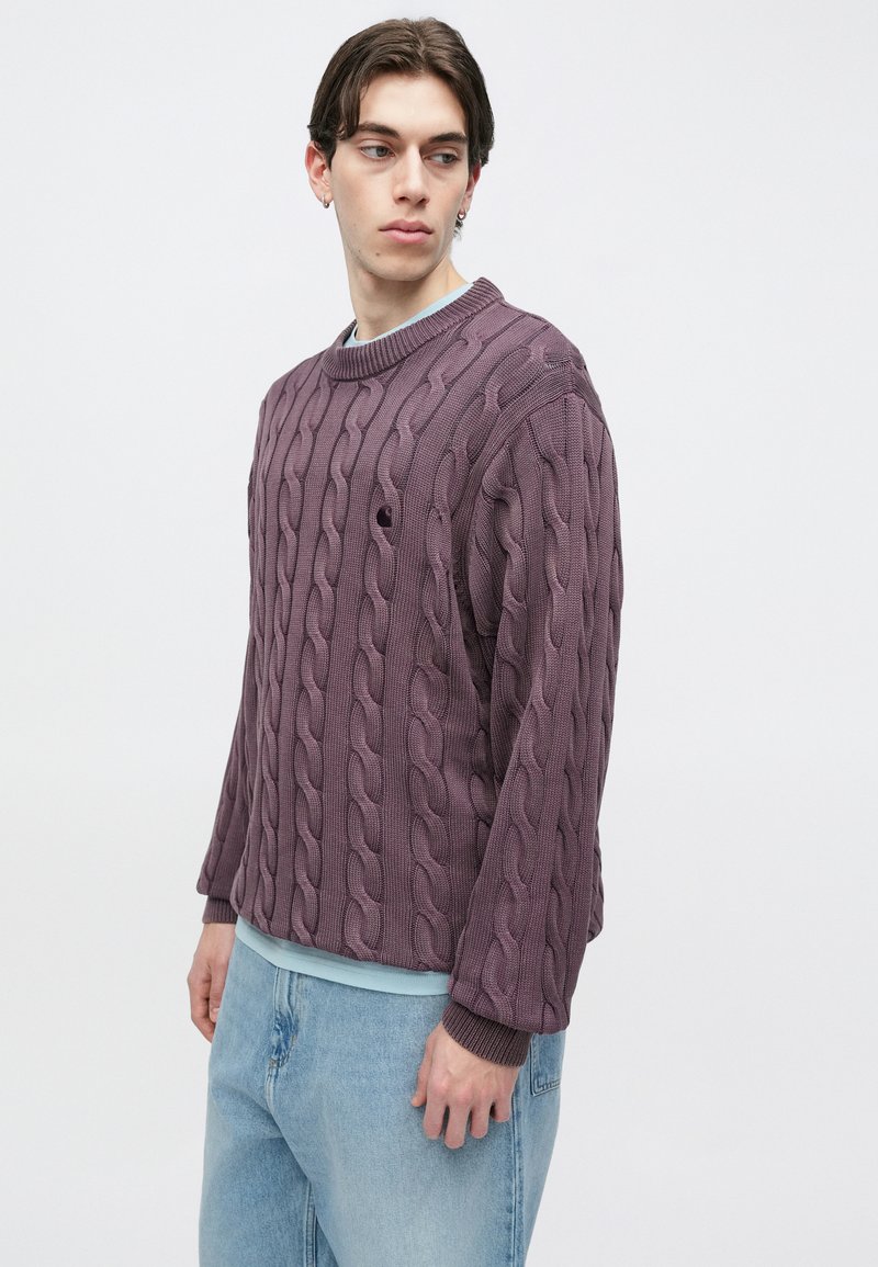 Strickpullover mit Zopfmuster in gedämpftem Lila, mit langen Ärmeln, rundem Ausschnitt und strukturiertem Rippenmuster. Kombiniert mit hellblauen Denim-Jeans.