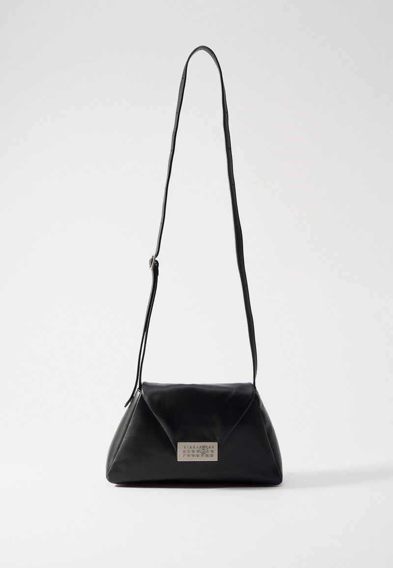 Musta nahkainen käsilaukku, jossa on kolmion muotoinen muotoilu, pitkä hihna ja suorakulmainen metallinen koristus etupuolella. Tasainen pinta.