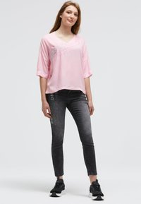 Woman standing with hands relaxed, wearing a loose pink T-shirt and faded black jeans with white text, paired with black sneakers.