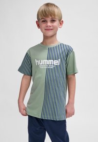 T-shirt à manches courtes en vert olive avec un panneau rayé vertical bleu marine. Présente le texte imprimé en blanc "hummel" et un slogan à l'avant.
