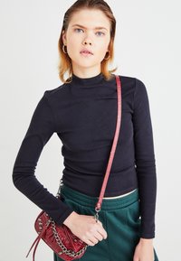 Marineblå langermet topp med høy hals, kombinert med grønne bukser med snøring. Holder en liten rød crossover-bag med kjedeaksenter.
