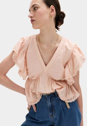 Kvinde med mørkt hår sat op i en knold iført en ferskenfarvet bluse med flæser og blondekanter, blå højtaljede jeans, guldkæde og abstrakte guldbørn.