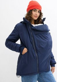 Giacca imbottita blu navy con una parte anteriore staccabile per il babywearing, caratterizzata da una chiusura con zip, grandi tasche e cappuccio. Texture liscia.