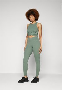 Oliven grønn idretts crop top med vridd front, kombinert med matchende høyt midje leggings. Sorte løpesko fullfører antrekket.