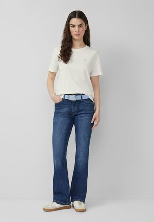 Vrouw met lang donker haar die een wit shirt met korte mouwen, een blauwe riem, donkerblauwe flare-jeans en witte sneakers draagt, staand tegen een grijze muur.